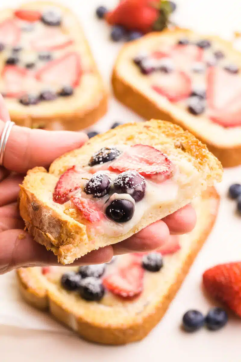 A hand holds a slice of air-fried custard toast with berries on top. There are more slices visible in the background, along with fresh berries.