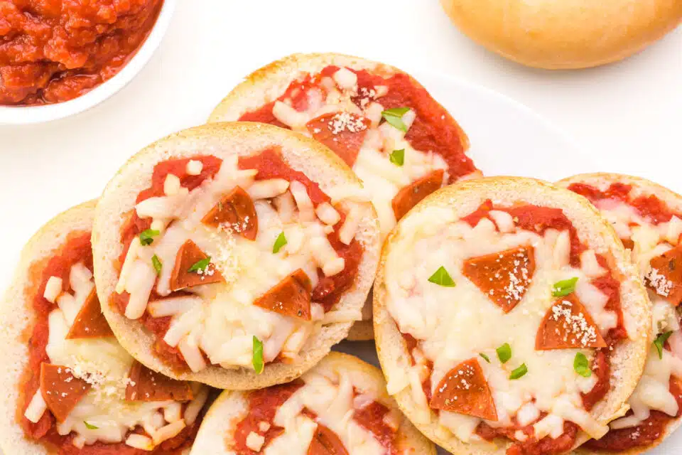 Looking down on pizza bagel bites on a plate. They sit next to a bowl of marinara sauce and a bagel, both of which are barely visible.
