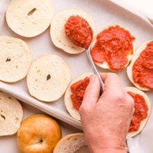A hand holds a spoon, spreading pizza sauce over mini bagels laid out on a baking pan.