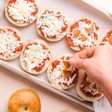 A hand puts vegan pepperoni pizza on mini bagels topped with pizza sauce and vegan mozzarella shreds. There are mini bagels sitting by the pan.
