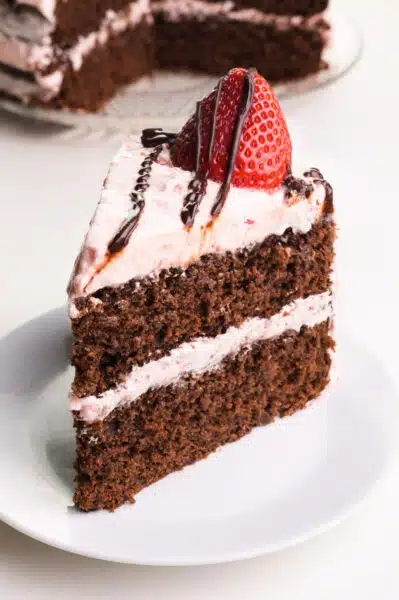 A slice of chocolate layer cake has strawberry frosting with a fresh strawberry and chocolate sauce on top.