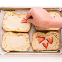A hand is adding sliced strawberries on top of pieces of bread topped with yogurt custard in a baking pan.