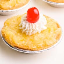 A pineapple tart has whipped cream on top and a maraschino cherry. Other tarts are barely visible in the background.