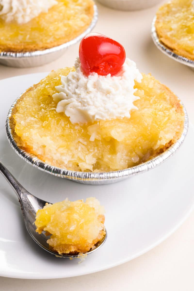 A bite sits on a spoon in front of a vegan pineapple tart. The tart has whipped cream on top with a cherry. There are other tarts barely visible in the background.