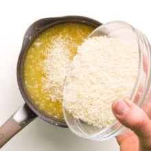 A hand holds a bowl of coconut flakes, pouring them into a saucepan with a pineapple mixture.