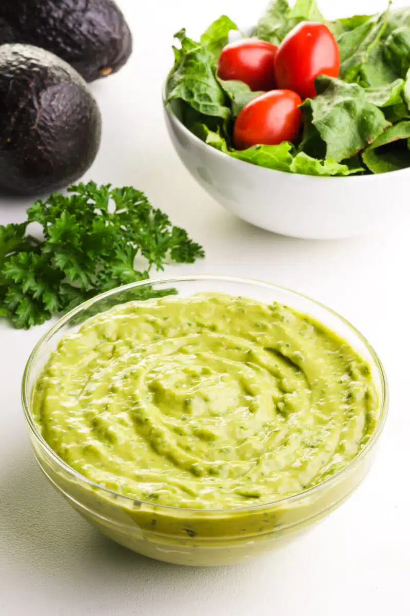 A bowl of green goddess dressing sits in front of a bowl of salad with tomatoes, two avocados, and fresh herbs.