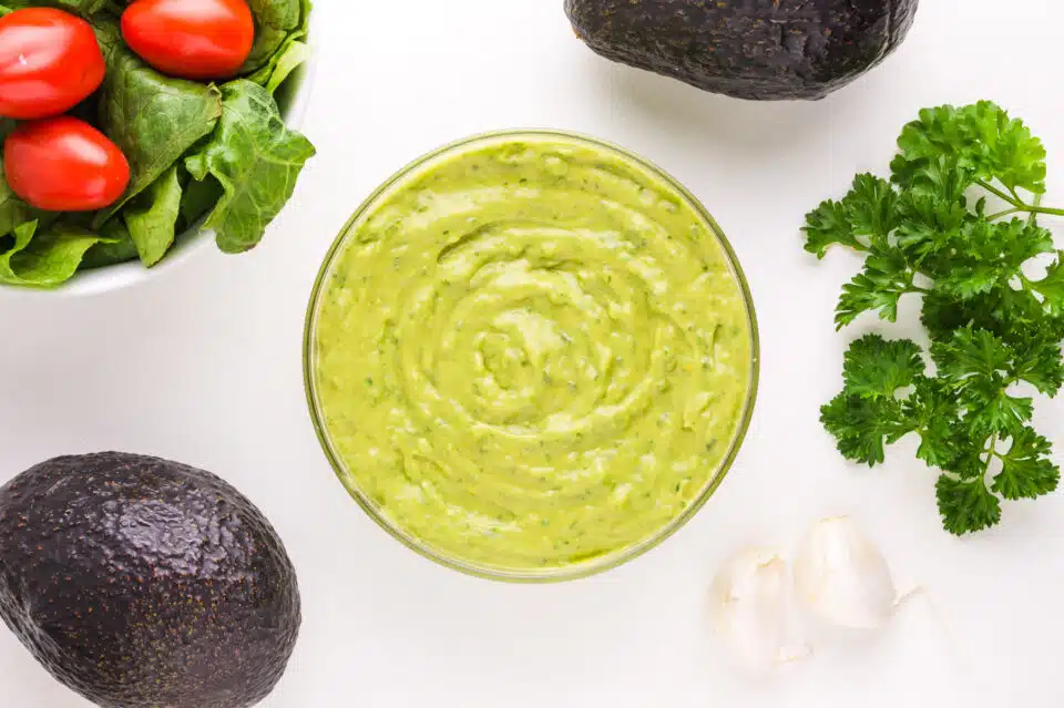 Looking down on avocado dressing surrounded by garlic, herbs, avocados, and a salad.