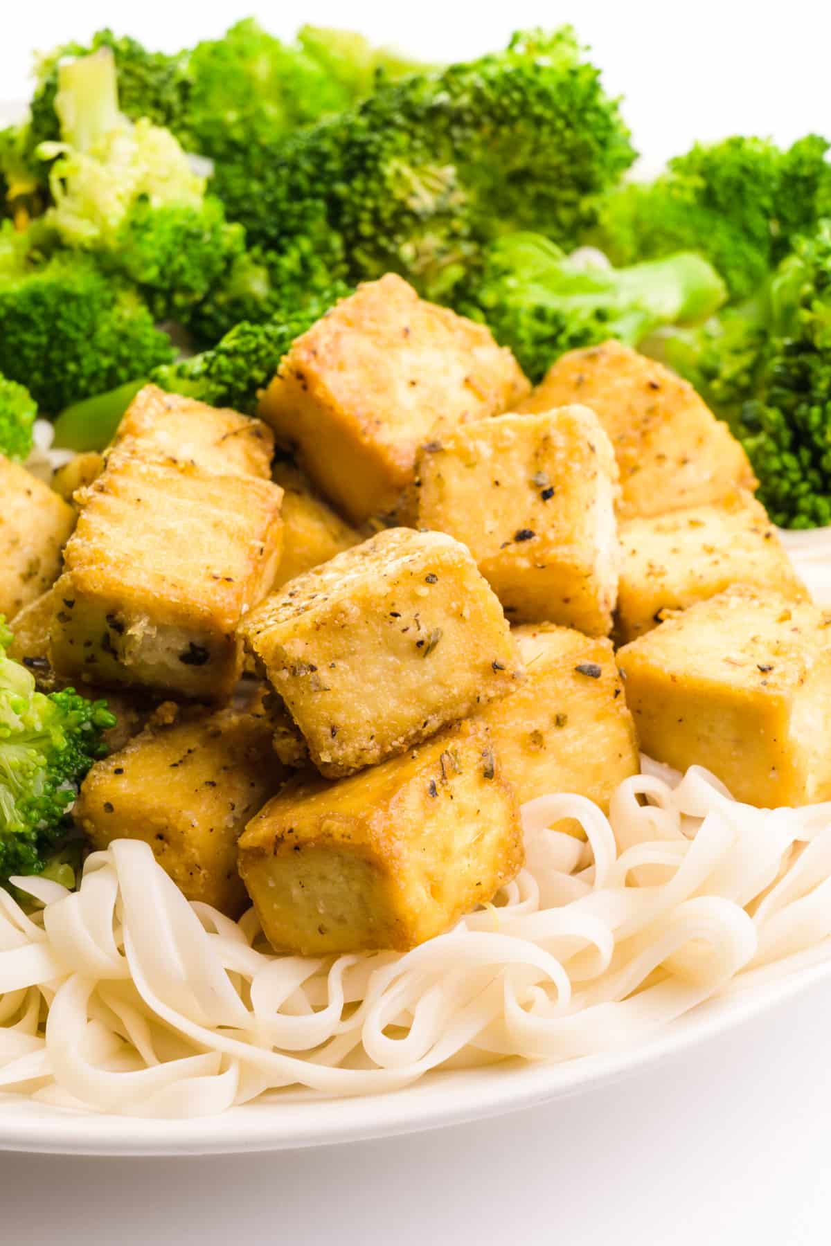 Savory baked tofu sits on a plate with noodles and steamed broccoli.