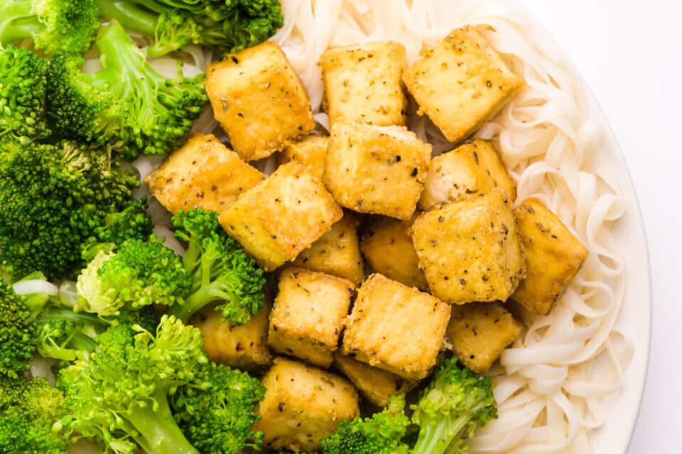Looking down on crispy tofu cubes on a plate with noodles and steamed broccoli.