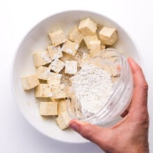 cornstarch is being spriled over tofu cubes in a bowl.