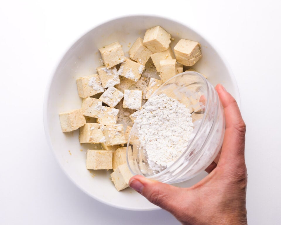 cornstarch is being sprinkled over tofu cubes in a bowl.