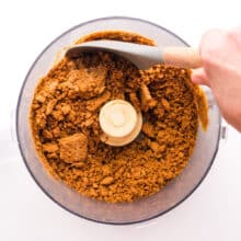 A hand holds a spatula, scraping down the sides of a food processor bowl with crumbled biscoff cookies in it.