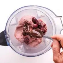 A hand drizzles spoonfuls of melted chocolate into a blender with a frozen cherry mixture.