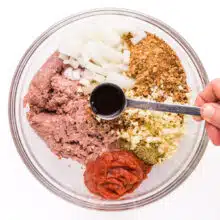 A hand holds a measuring spoon of sauce, pouring it over ingredients such as breadcrumbs and onions in a bowl.