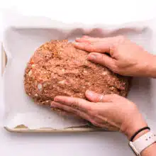 Two hands mold meatloaf mixture into an oblong shape on a baking pan lined with parchment paper.