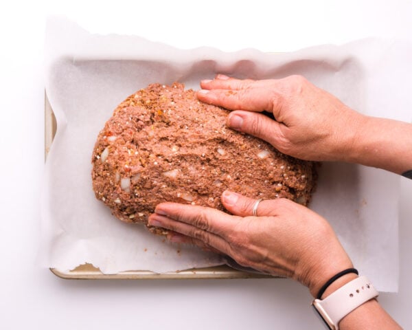 Two hands mold meatloaf mixture into an oblong shape on a baking pan lined with parchment paper.
