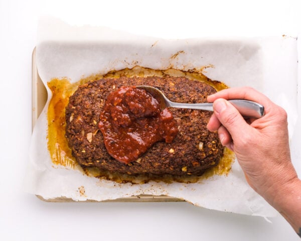 A hand holds a spoon, pouring tomato sauce over a baked meatloaf in a pan.
