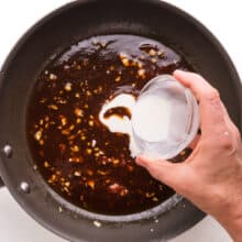 A hand holds a scowl bowl of cornstarch slurry, pouring it into a skillet with a sauce mixture.