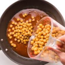 A hand holds a glass measuring cup full of chickpeas, pouring them into a skillet with sauce.