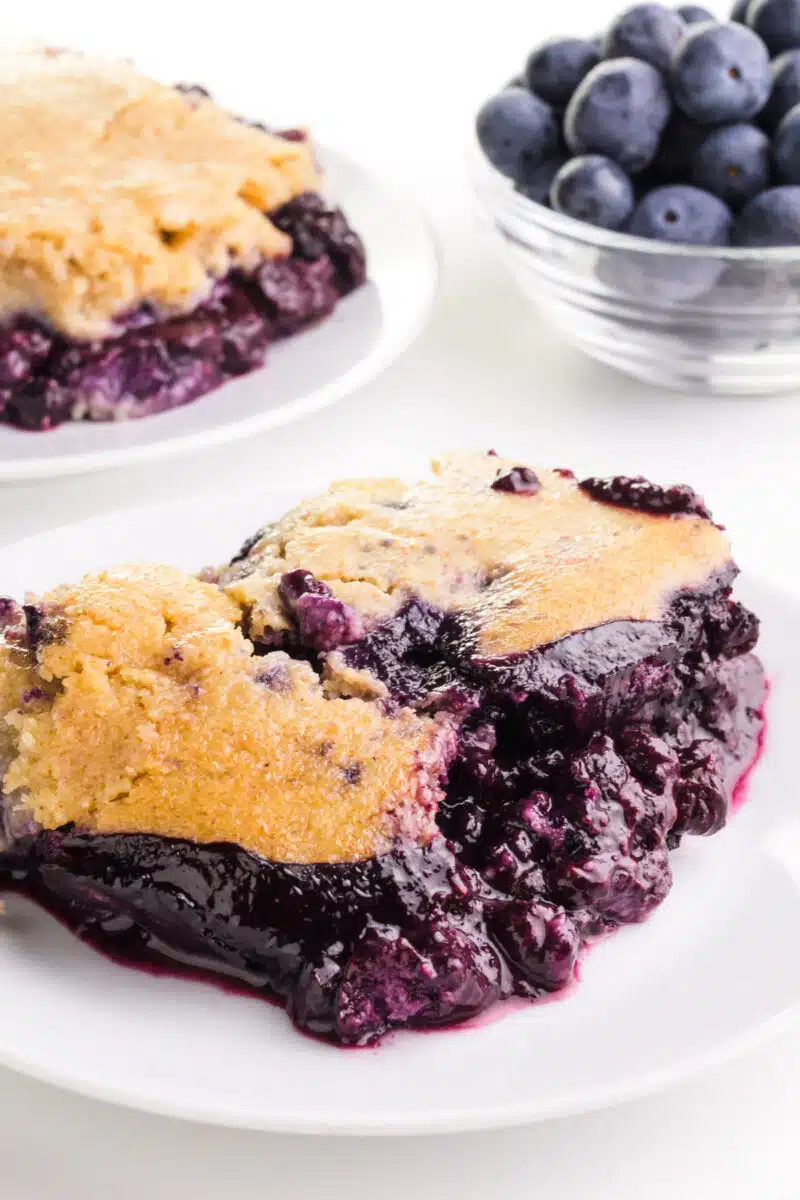 A slice of blueberry cobbler sits on a plate. There is another plate of the dessert and a bowl of fresh blueberries in the background.