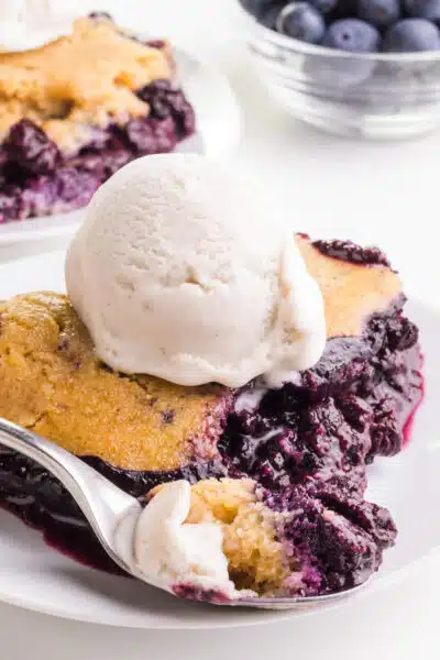 A slice of vegan blueberry cobbler has ice cream on top and has a spoonful resting in front. There is another slice and a bowl of blueberries in the back.