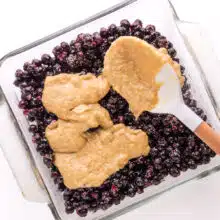 Batter is being spooned over a blueberry mixture in a baking pan.