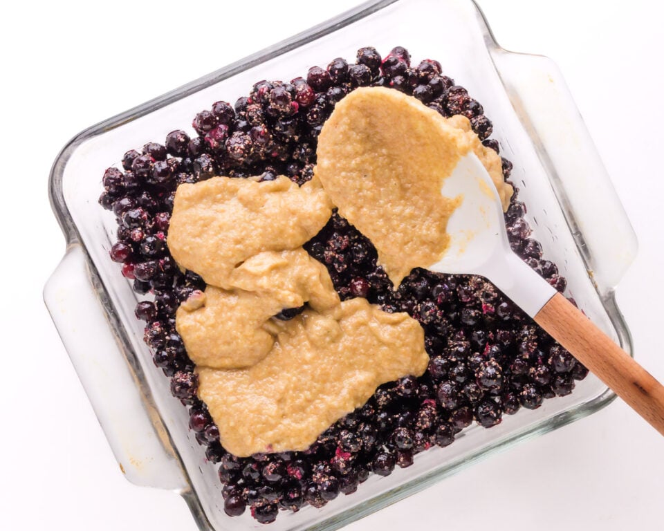 Batter is being spooned over a blueberry mixture in a baking pan.