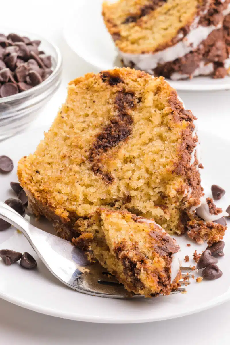 A slice of vegan chocolate coffee cake sits on a plate with a fork with a bite in front of it. There is a bowl of chocolate chips and another slice in the background.