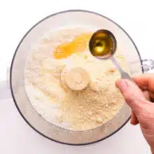 A hand holds a measuring spoon pouring syrup into a food processor with coconut flakes.