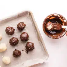 A hand holds a fork, dipping coconut balls in melted chocolate. There are more coconut balls on a pan, some are dipped others are not.