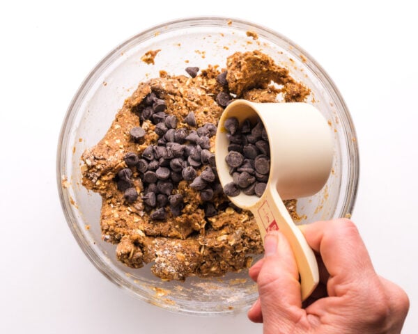 A hand holds a measuring cup of chocolate chips, pouring it into a bowl with cookie dough.