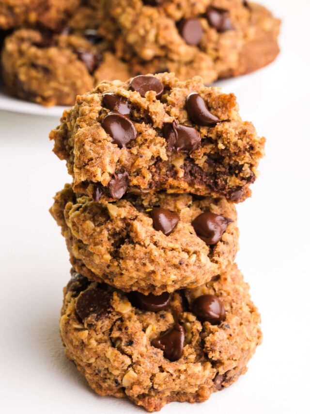 A stack of vegan coffee cookies shows the top one with a bite taken out. There is a plate with more cookies in the background.