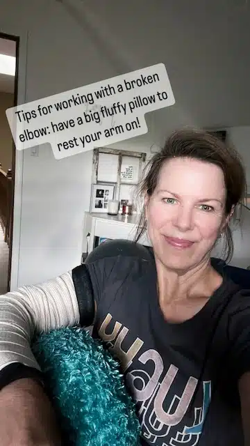 Marly sits in an office chair with a blue pillow. The text reads, Tips for working with a broken elbow: have a big fluffy pillow to rest your arm on!