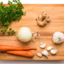 Ingredients like garlic, onions, ginger, carrots, and parsley are on a cutting board.