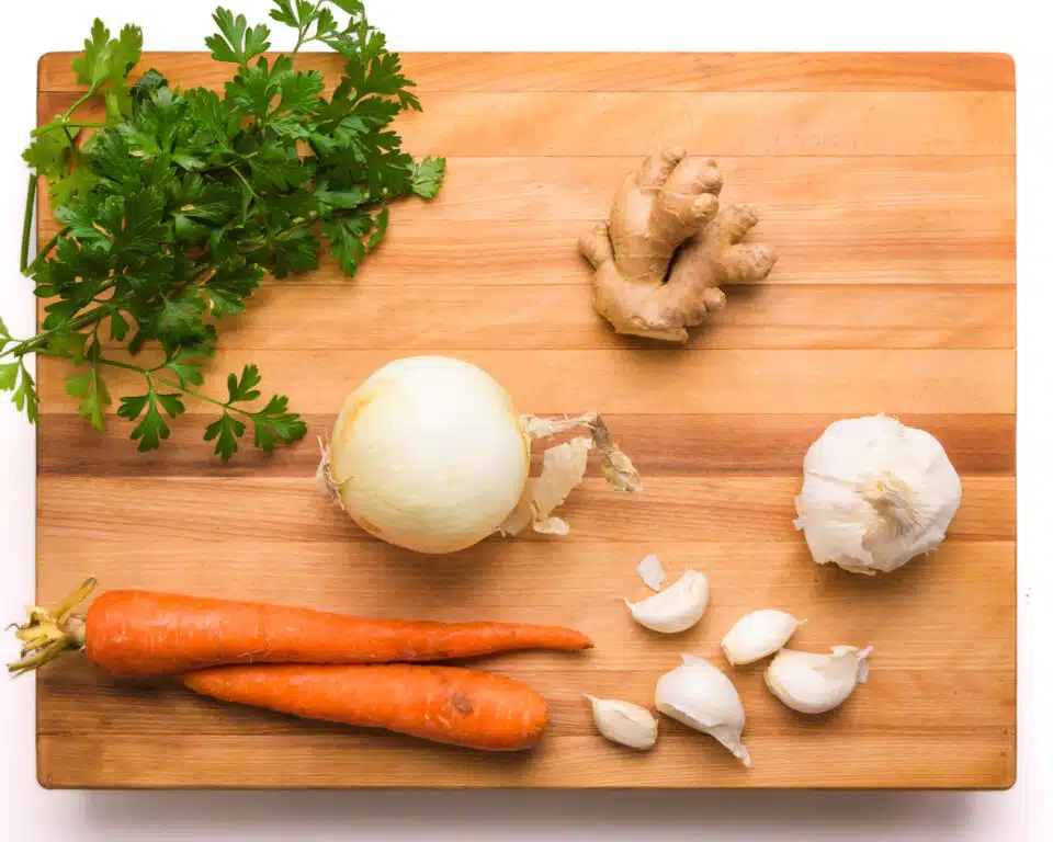 Ingredients like garlic, onions, ginger, carrots, and parsley are on a cutting board.