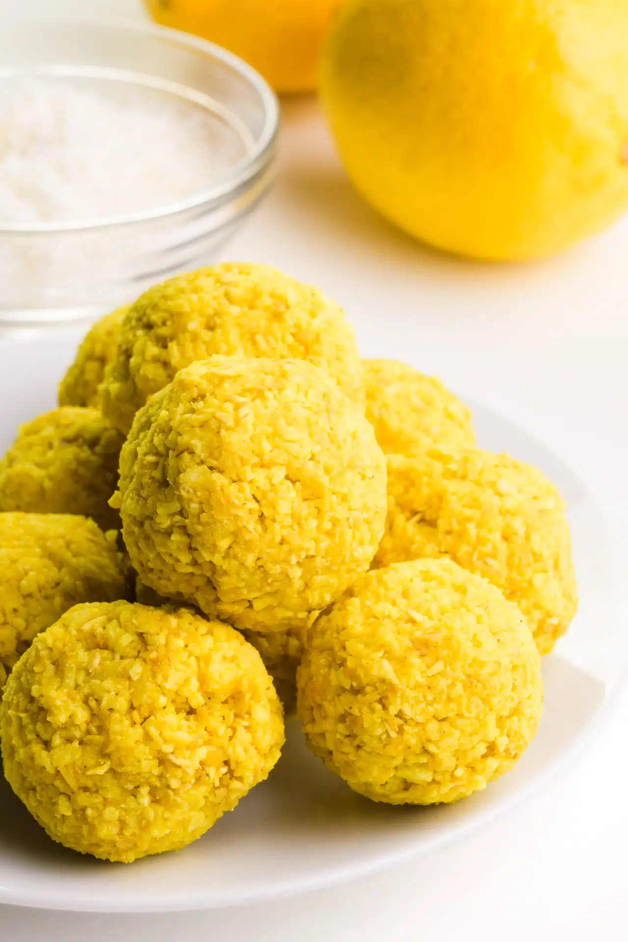 A stack of lemon energy balls are on a plate, sitting in front of a bowl of coconut flakes and fresh lemons.
