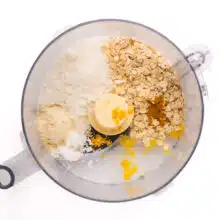 Looking down on ingredients in a food processor bowl, such as oats, lemon zest, coconut flakes, and more.