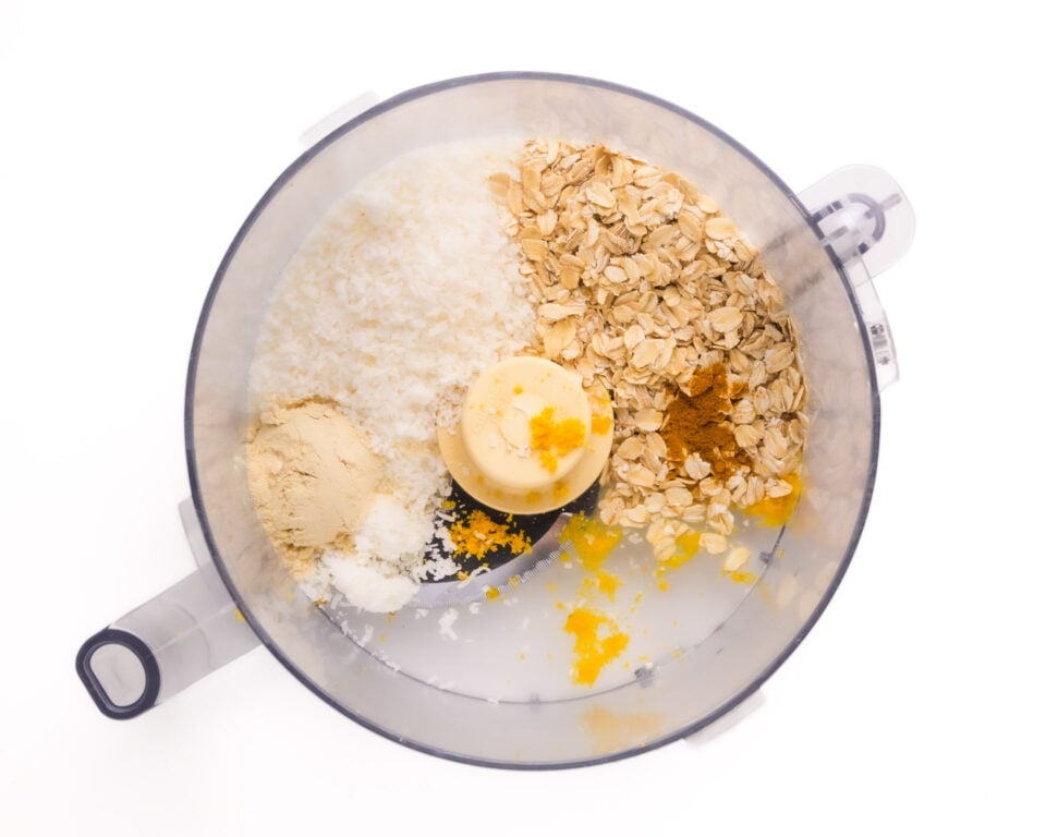 Looking down on ingredients in a food processor bowl, such as oats, lemon zest, coconut flakes, and more.