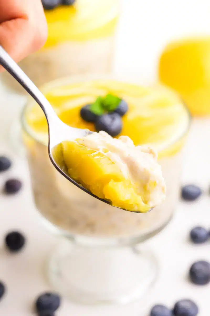 A hand holds a spoonful of lemon overnight oats. It hovers in front of more overnight oats, blueberries, and a lemon in the background.