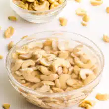 A bowl of cashews are soaking in water. There is a bowl of raw cashews in the background and more strewn across the table.