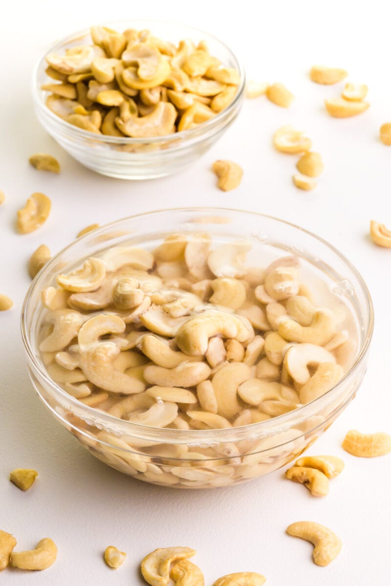 A bowl of cashews are soaking in water. There is a bowl of raw cashews in the background and more strewn across the table.