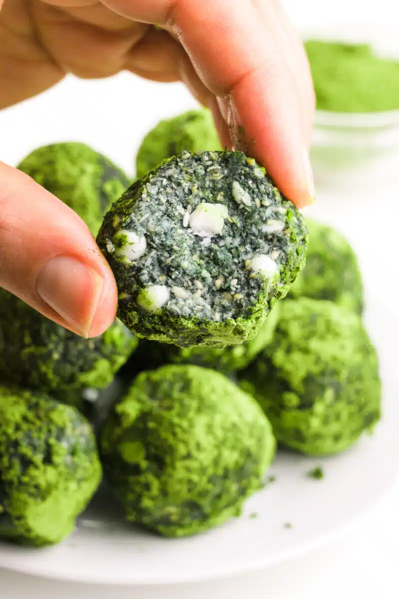 A hand holds an energy ball with a bite taken out, revealing white chocolate chips inside. There is a plate with more energy balls and a bowl of matcha powder in the background.