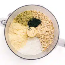 Looking down on ingredients in a food processor, including oats, coconut flakes, and spirulina.