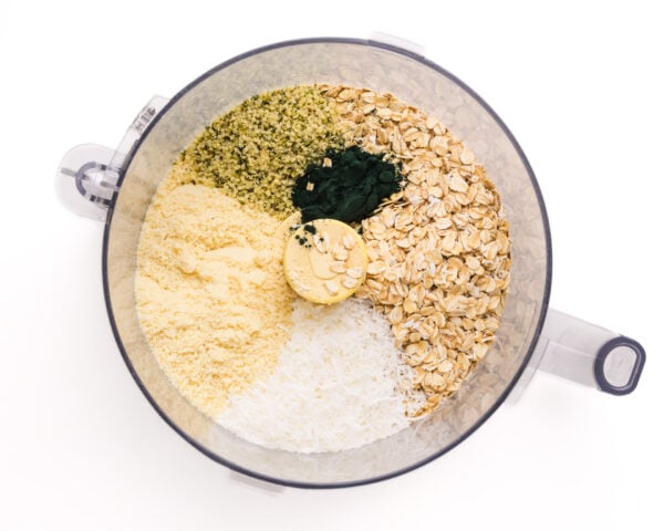 Looking down on ingredients in a food processor, including oats, coconut flakes, and spirulina.