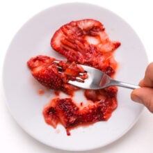 A hand holds a fork, mashing strawberries on a plate.