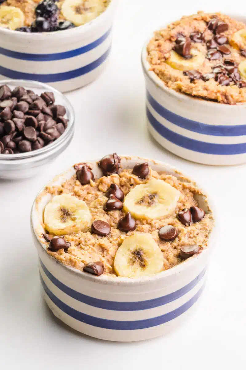 Three bowls of baked oats have chocolate chips and banana slices on top. They sit next to a bowl of chocolate chips.