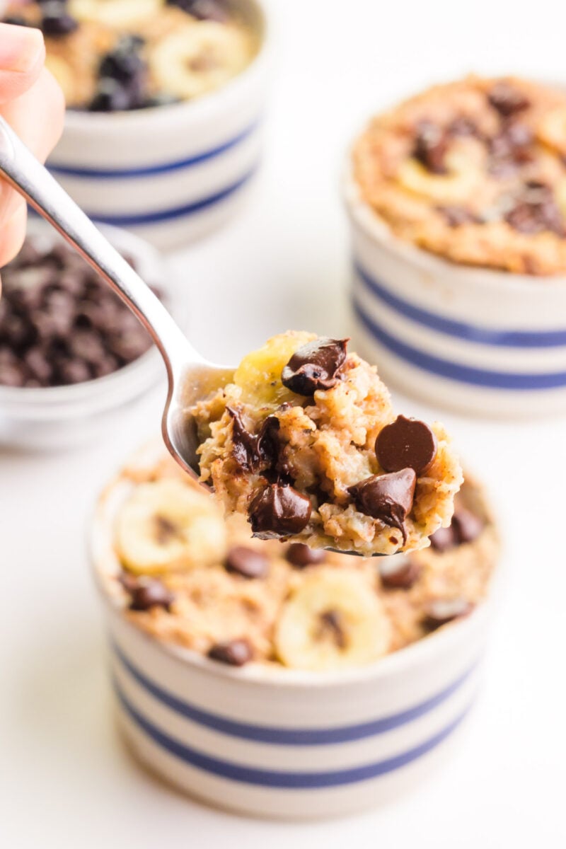 A hand holds a spoon with a bite of baked oats on it. There are bowls with baked oats in the background.