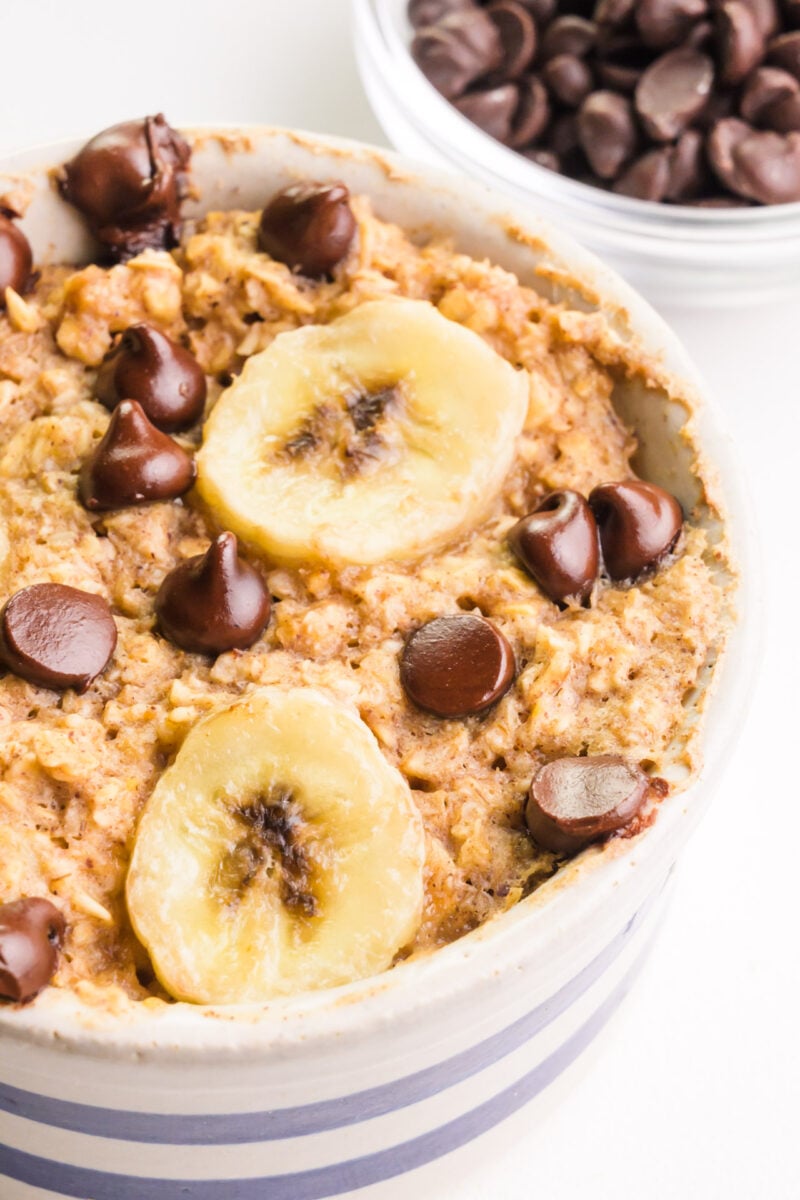 A bowl of vegan baked oats has chocolate chips and banana slices on top. There is a bowl of chocolate chips in the background.
