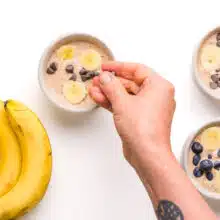 A hand drops toppings onto batter in a ramekin. There are two more batter-filled ramekins beside it along with whole bananas.