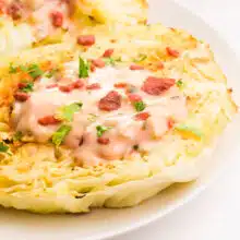 Cabbage steaks are sitting on a plate. They are topped with dressing and veggie bacon bits.